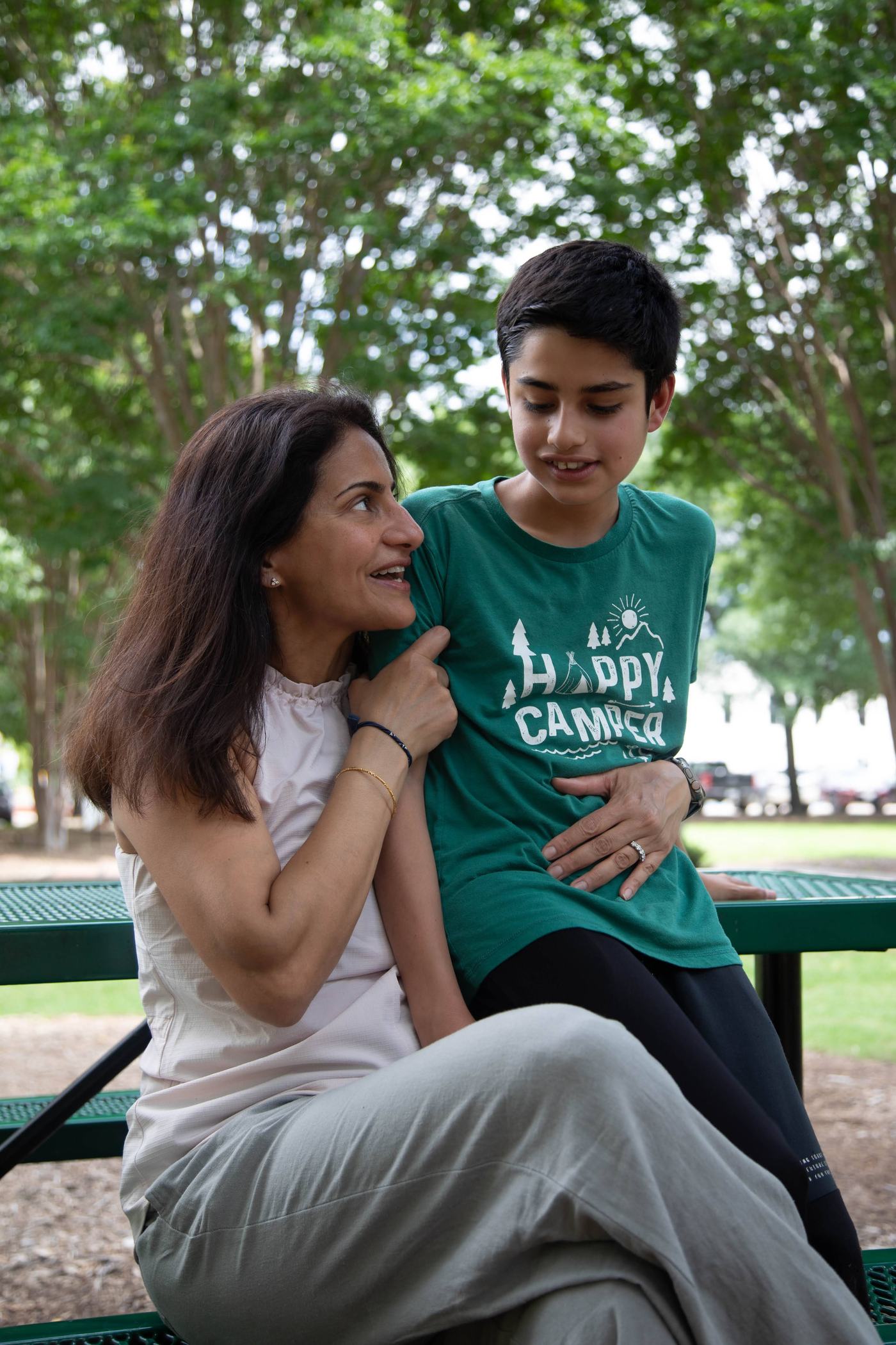 A mother laughs with her teenage son on a park bench.